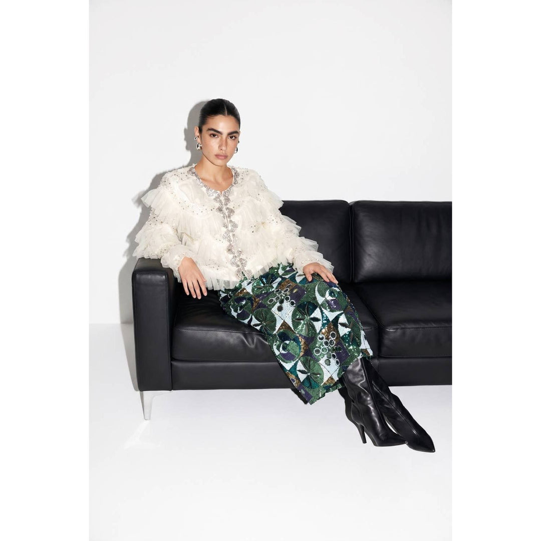 A woman wearing a green sequin embellished skirt paired with a white ruffled top and black high-heeled boots