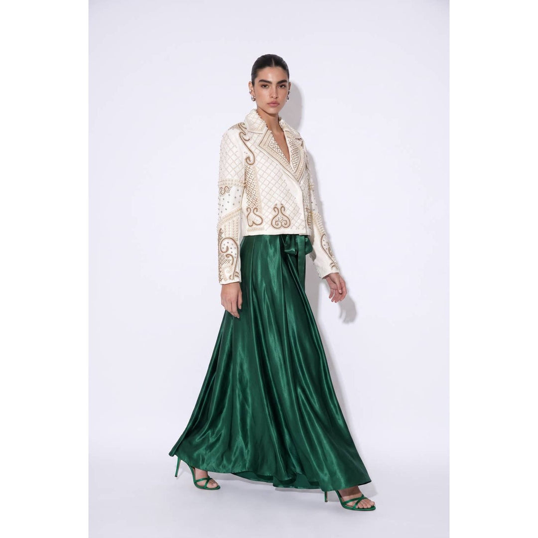A woman wears an emerald green glossy satin maxi skirt with an ivory jacket featuring gold embroidery