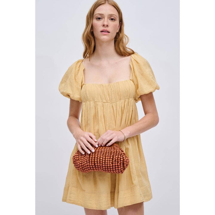 Model holding The Pearl Pagoda Lydia beaded teak wood clutch with a gold chain strap, wearing a yellow mini dress.