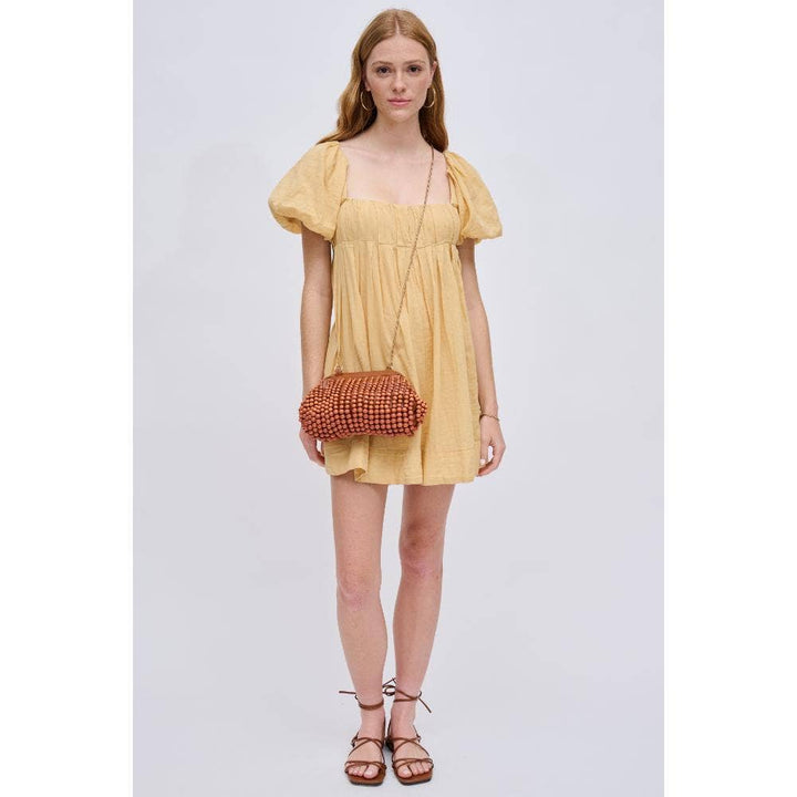 The Pearl Pagoda Lydia beaded clutch in teak wood held by a model wearing a yellow puff-sleeve dress and gold chain.